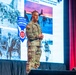 A U.S. Army officer in full camouflage uniform (OCP/MultiCam) stands on a stage delivering a presentation. The backdrop displays a large projection screen showing the '11th Airborne Division - Arctic Airborne' insignia alongside historical and modern military photographs. The setting appears to be a formal military conference or briefing event.
