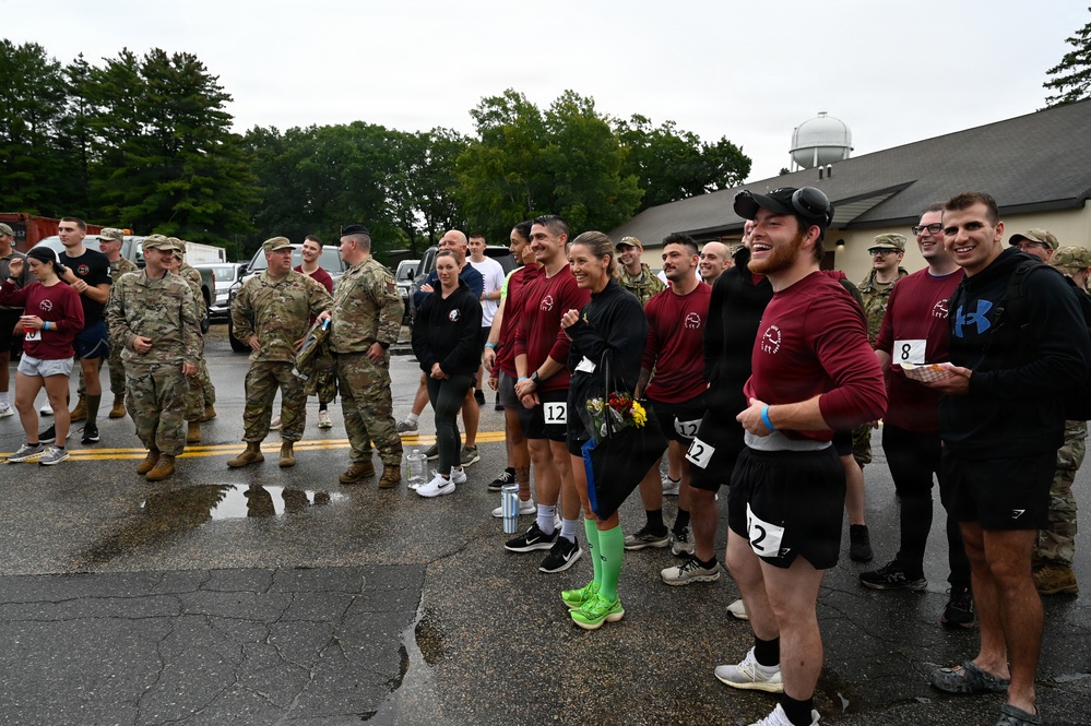 A group of military personnel and civilian athletes are gathered on a wet parking lot, likely after completing a race or athletic competition. Participants wearing matching maroon shirts with race bib numbers (visible numbers include 2, 8, and 12) are standing together smiling and laughing, while uniformed soldiers in camouflage (OCP pattern) stand nearby. The overcast, rainy conditions and wet pavement suggest the event took place in inclement weather.