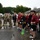 157th Airmen Compete in 2026 Pease Warrior Challenge, Demonstrate Physical Prowess A group of military personnel and civilian athletes are gathered on a wet parking lot, likely after completing a race or athletic competition. Participants wearing matching maroon shirts with race bib numbers (visible numbers include 2, 8, and 12) are standing together smiling and laughing, while uniformed soldiers in camouflage (OCP pattern) stand nearby. The overcast, rainy conditions and wet pavement suggest the event took place in inclement weather.