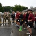A group of military personnel and civilian athletes are gathered on a wet parking lot, likely after completing a race or athletic competition. Participants wearing matching maroon shirts with race bib numbers (visible numbers include 2, 8, and 12) are standing together smiling and laughing, while uniformed soldiers in camouflage (OCP pattern) stand nearby. The overcast, rainy conditions and wet pavement suggest the event took place in inclement weather.