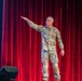 A U.S. Army officer in OCP (Operational Camouflage Pattern) uniform stands on a stage with a red curtain backdrop, gesturing with an outstretched arm toward the left. The individual appears to be presenting or speaking to an audience, likely at a formal military briefing or event. A projection screen is partially visible on the left side of the stage.