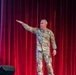 A U.S. Army soldier in OCP (Operational Camouflage Pattern) uniform is standing on a stage in front of a red curtain, gesturing with an outstretched arm toward the left. The individual appears to be presenting or speaking to an audience, with a projection screen partially visible on the left side of the stage. The setting suggests a formal military briefing, ceremony, or educational event held in an auditorium or theater.