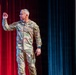 A U.S. Army soldier, identified by the name tape 'HANSON' on his uniform, stands on a stage delivering a presentation or speech. He is wearing MultiCam (OCP) camouflage uniform with a U.S. Army tape, rank insignia, and a shoulder patch. His right hand is raised in a gesturing motion, suggesting active engagement with an audience.