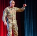A U.S. Army soldier in OCP (Operational Camouflage Pattern) uniform is standing on a stage, appearing to give a presentation or speech. The name tape reads 'HANSON' and the uniform displays 'U.S. ARMY' tape, a rank insignia, and a shoulder patch. The soldier is gesturing with a raised fist toward the audience against a backdrop of red and teal stage curtains.