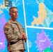 A U.S. Army soldier in MultiCam/OCP camouflage uniform is presenting or briefing in front of a large digital display screen showing military operational maps with color-coded zones. The name tape on his uniform reads 'COGBILL' and he wears a U.S. flag patch on his left shoulder. The maps behind him appear to depict tactical military planning areas labeled with terms such as 'Corps Rear' and 'Corps Close.'