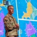 A U.S. Army officer in camouflage uniform stands before a large tactical operations display screen showing color-coded military maps. The officer appears to be delivering a briefing or presentation, with map panels visible behind him showing areas labeled 'Corps Rear' and 'Corps Group'. The display uses multiple colors (purple, yellow, green, blue) to denote different operational zones.