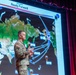 A U.S. Army officer in OCP (Operational Camouflage Pattern) uniform is presenting a briefing on stage in front of a large projected map slide titled 'Ready Forward' marked as 'UNCLASSIFIED'. The slide displays a world map with multiple arrows and military exercise names indicating global deployment or operational reach. The setting appears to be a formal military briefing or conference venue.