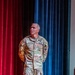 A U.S. Army soldier named Hanson, identifiable by his name tape and branch tape, stands on a stage in front of red and blue curtains. He is wearing the Army Combat Uniform (ACU) in OCP (Operational Camouflage Pattern) and appears to be speaking or presenting at a formal military event, indicated by a lapel microphone clipped to his uniform. His rank insignia on the chest suggests a senior non-commissioned officer.