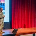 Two U.S. Army soldiers in camouflage uniforms are on a stage, with one appearing to give a presentation or briefing. Behind them is a projection screen displaying the insignia and imagery of the 11th Airborne Division ('Arctic Airborne'). The setting appears to be a military auditorium or theater with red curtains.