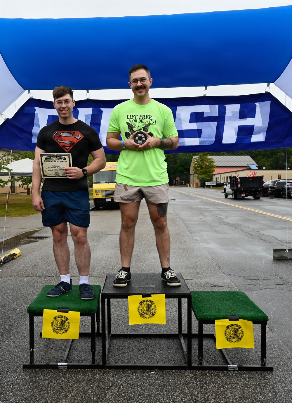 Two men stand on a competition podium under a blue finish-line banner, with the taller man in a neon green 'Lift Free or Die' shirt occupying the 1st place position holding a medal/award, and the shorter man in a Superman shirt standing on the 2nd place platform holding a certificate. The podium platforms are covered in green turf and labeled with yellow signs reading '1st Place Winner,' '2nd Place Runner Up,' and '3rd Place.' The setting appears to be an outdoor fitness or strength competition held in a parking lot or base area.