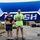 157th Airmen Compete in 2026 Pease Warrior Challenge, Demonstrate Physical Prowess Two men stand on a competition podium under a blue finish-line banner, with the taller man in a neon green 'Lift Free or Die' shirt occupying the 1st place position holding a medal/award, and the shorter man in a Superman shirt standing on the 2nd place platform holding a certificate. The podium platforms are covered in green turf and labeled with yellow signs reading '1st Place Winner,' '2nd Place Runner Up,' and '3rd Place.' The setting appears to be an outdoor fitness or strength competition held in a parking lot or base area.