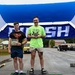 Two men stand on a competition podium under a blue finish-line banner, with the taller man in a neon green 'Lift Free or Die' shirt occupying the 1st place position holding a medal/award, and the shorter man in a Superman shirt standing on the 2nd place platform holding a certificate. The podium platforms are covered in green turf and labeled with yellow signs reading '1st Place Winner,' '2nd Place Runner Up,' and '3rd Place.' The setting appears to be an outdoor fitness or strength competition held in a parking lot or base area.