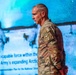 A U.S. Army soldier in OCP (Operational Camouflage Pattern) uniform stands in profile view on what appears to be a stage or presentation area. The background displays a projected presentation slide referencing Arctic capabilities and Army Arctic strategy. The soldier wears shoulder patches indicating Ranger, Arctic, and Airborne qualifications.