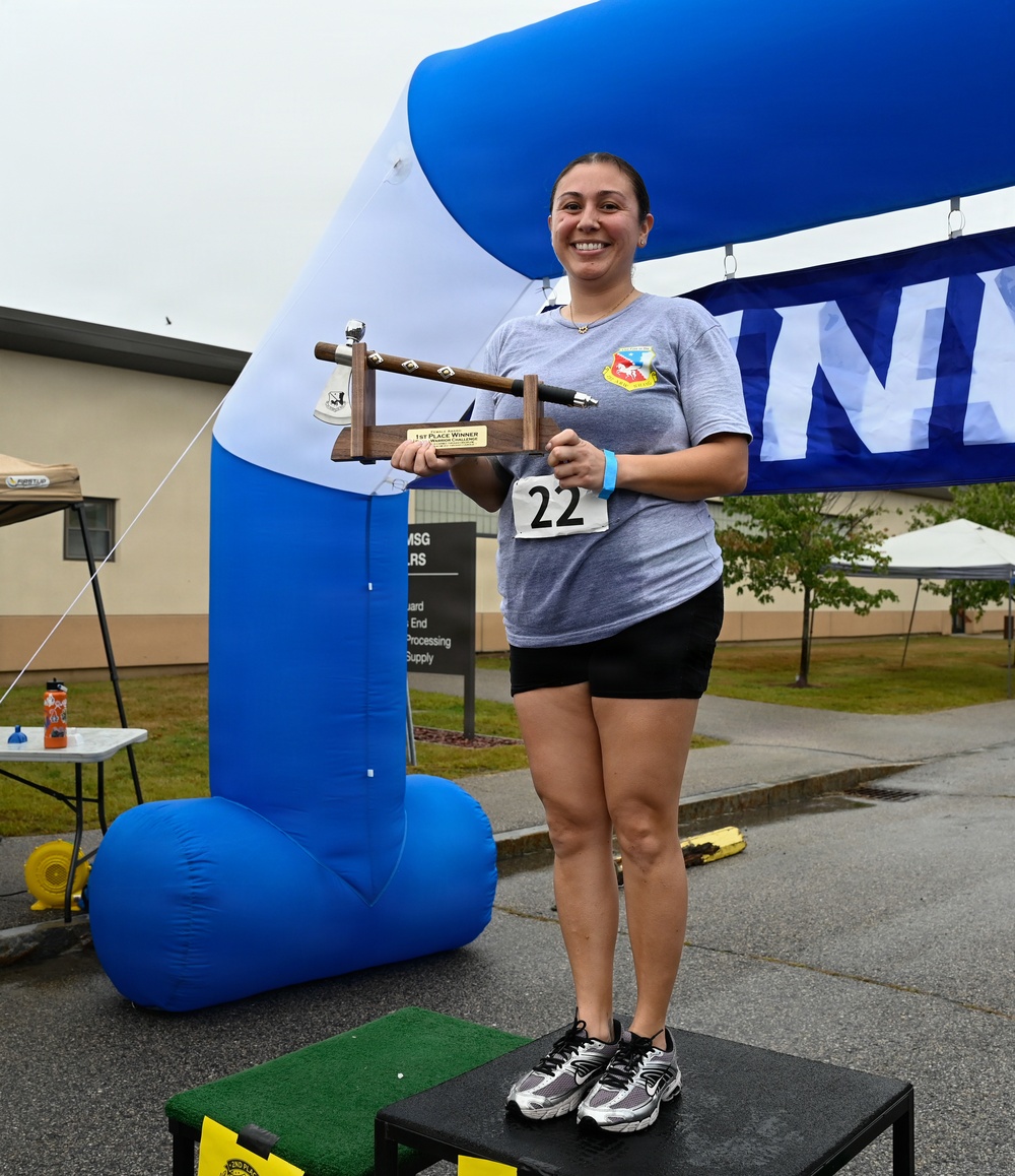 A smiling woman wearing race bib number 22 stands on a winner's podium holding a trophy with a '1st Place Winner' plaque. She is positioned under a blue and white inflatable finish line arch at what appears to be a military base running event. Her gray t-shirt displays a military unit patch/emblem.