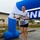 157th Airmen Compete in 2026 Pease Warrior Challenge, Demonstrate Physical Prowess A smiling woman wearing race bib number 22 stands on a winner's podium holding a trophy with a '1st Place Winner' plaque. She is positioned under a blue and white inflatable finish line arch at what appears to be a military base running event. Her gray t-shirt displays a military unit patch/emblem.