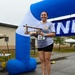 A smiling woman wearing race bib number 22 stands on a winner's podium holding a trophy with a '1st Place Winner' plaque. She is positioned under a blue and white inflatable finish line arch at what appears to be a military base running event. Her gray t-shirt displays a military unit patch/emblem.
