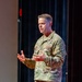 A U.S. Army officer in OCP (Operational Camouflage Pattern) uniform is speaking or presenting on a stage, gesturing with both hands. His name tape reads 'COGBILL' and he wears a rank insignia consistent with a general officer (one star visible). He has multiple shoulder patches including Ranger, Arctic, and Airborne tabs.