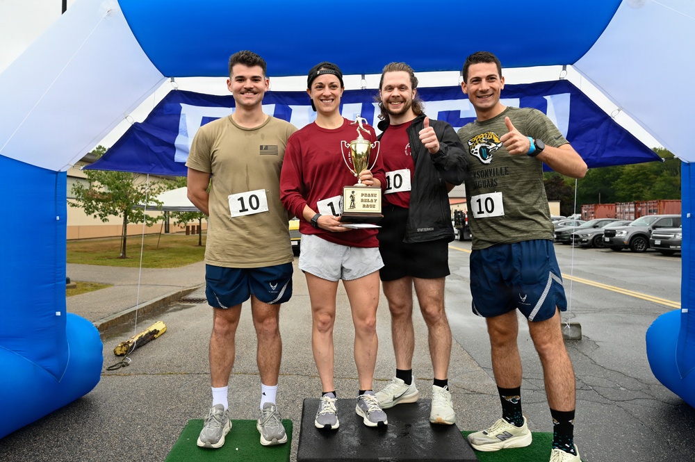 Four individuals pose together under a blue inflatable arch at what appears to be a relay race finish line. They are all wearing race bib number '10' and one person is holding a trophy labeled 'Pease Relay Race,' indicating they are the winning team. The group appears celebratory, with smiles and thumbs-up gestures.