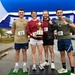 Four individuals pose together under a blue inflatable arch at what appears to be a relay race finish line. They are all wearing race bib number '10' and one person is holding a trophy labeled 'Pease Relay Race,' indicating they are the winning team. The group appears celebratory, with smiles and thumbs-up gestures.