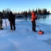 A group of people are standing on a frozen lake or river in what appears to be a subarctic or boreal environment. One person in a red jacket is operating what appears to be ground-penetrating radar (GPR) or a similar geophysical survey instrument with a red sensor unit being dragged across the ice. Other individuals, some in military-style clothing, stand nearby observing, with an ice auger visible on the left side.