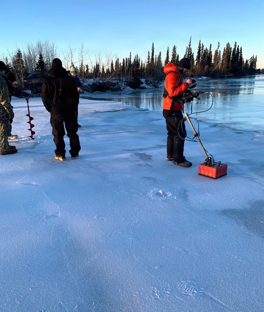 A group of people are standing on a frozen lake or river in what appears to be a subarctic or boreal environment. One person in a red jacket is operating what appears to be ground-penetrating radar (GPR) or a similar geophysical survey instrument with a red sensor unit being dragged across the ice. Other individuals, some in military-style clothing, stand nearby observing, with an ice auger visible on the left side.