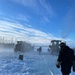 Workers in cold-weather gear operate on a frozen surface alongside heavy tracked and wheeled machinery, with visible steam or mist rising from the ice. A hose or cable runs along the ice surface toward the equipment in the background. The scene appears to depict ice road construction or maintenance operations in a subarctic or arctic environment.