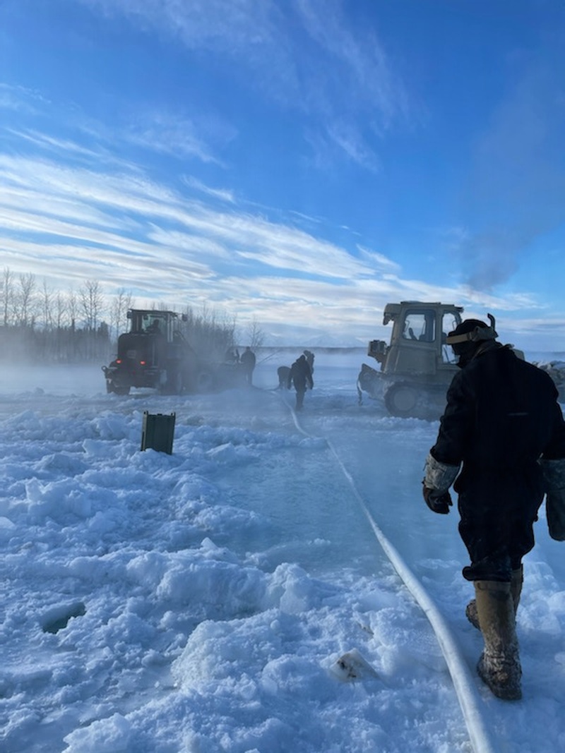 Workers in cold-weather gear operate on a frozen surface alongside heavy tracked and wheeled machinery, with visible steam or mist rising from the ice. A hose or cable runs along the ice surface toward the equipment in the background. The scene appears to depict ice road construction or maintenance operations in a subarctic or arctic environment.
