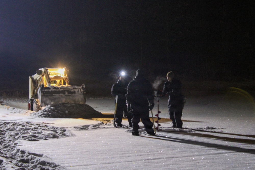 A nighttime scene showing three individuals dressed in dark winter clothing standing on a snow-covered surface, with one person wearing a headlamp. They appear to be operating an ice auger, likely drilling into a frozen body of water or snow-covered ground. A yellow construction vehicle, possibly a front-end loader or backhoe, is illuminated in the background to the left.