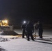 A nighttime scene showing three individuals dressed in dark winter clothing standing on a snow-covered surface, with one person wearing a headlamp. They appear to be operating an ice auger, likely drilling into a frozen body of water or snow-covered ground. A yellow construction vehicle, possibly a front-end loader or backhoe, is illuminated in the background to the left.
