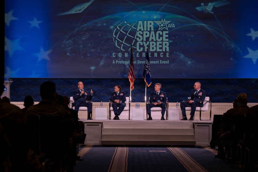 Four military officers in US Air Force dress uniforms are seated on a stage panel at the AFA Air, Space &amp; Cyber Conference, a professional development event. The leftmost officer is speaking into a microphone while the others listen. An audience is visible in silhouette in the foreground.