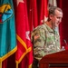 A U.S. Army soldier in Operational Camouflage Pattern (OCP) uniform is speaking at a wooden podium during what appears to be a formal military ceremony or briefing. The soldier's name tape reads 'ARMSTRONG' and visible patches include a U.S. flag, a Ranger tab, and a unit patch. Multiple military flags and guidons are displayed on a stage backdrop with red curtains.