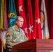 A U.S. Army Colonel named Armstrong is speaking at a wooden podium during a formal military ceremony or briefing. He is wearing the Army Combat Uniform (ACU) in Operational Camouflage Pattern (OCP) with visible name tape, branch tape, U.S. flag patch, and a Ranger tab. Multiple military flags and the U.S. flag are displayed on a red-curtained stage behind him.