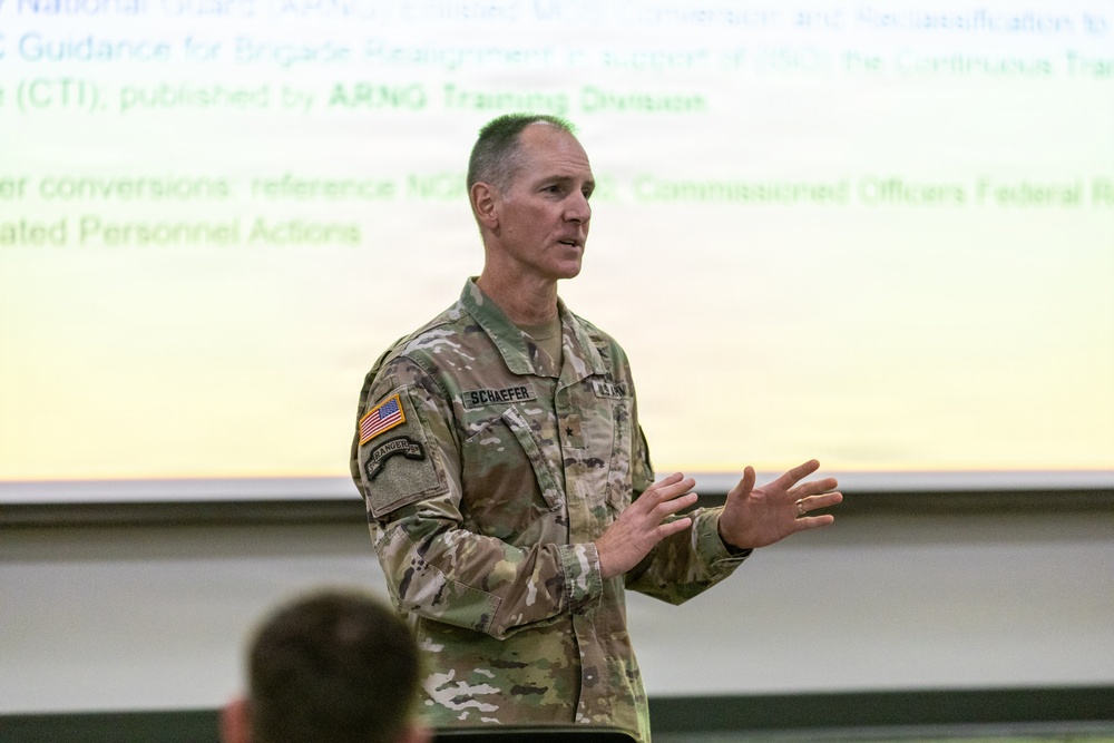 A U.S. Army officer identified by name tape as 'SCHAEFER' is delivering a briefing or presentation in what appears to be a military classroom or conference room. He is wearing the Army Combat Uniform (ACU) in MultiCam/OCP pattern with a single star insignia indicating the rank of Brigadier General. A projected presentation slide behind him references ARNG (Army National Guard) Training Division and Brigade Realignment topics.