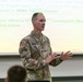 A U.S. Army officer identified by name tape as 'SCHAEFER' is delivering a briefing or presentation in what appears to be a military classroom or conference room. He is wearing the Army Combat Uniform (ACU) in MultiCam/OCP pattern with a single star insignia indicating the rank of Brigadier General. A projected presentation slide behind him references ARNG (Army National Guard) Training Division and Brigade Realignment topics.