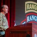 A U.S. Army soldier in camouflage uniform is speaking at a wooden podium during what appears to be a Ranger Regiment event or ceremony. The background features projected 'RANGER' tab insignia logos, and a red curtain is visible to the left. The setting appears to be an auditorium or formal presentation hall.