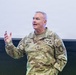 A U.S. Army officer identified by name tape as 'HOPKINS' is speaking or presenting, gesturing with his right hand. He is wearing an OCP (Operational Camouflage Pattern) uniform with visible rank insignia of a Major General (two stars) and multiple qualification badges. The setting appears to be an indoor briefing or lecture environment.