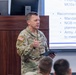 A U.S. Army Colonel (identifiable by eagle rank insignia) named TOOLEY is delivering a briefing or presentation in what appears to be a military conference or classroom setting. He is wearing Army Combat Uniform (ACU) with an American flag patch, Airborne tab, and 82nd Airborne Division patch visible on his sleeve. The presentation behind him references military topics including MOSs (Military Occupational Specialties), NGB (National Guard Bureau), and transformation recommendations.