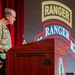 A U.S. Army soldier in camouflage uniform stands at a wooden podium delivering a speech at what appears to be a Ranger Regiment event or ceremony. The backdrop prominently displays 'RANGER' insignia logos, including what appears to be the 75th Ranger Regiment tab. Additional uniformed personnel are seated in the background to the right.