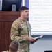 A U.S. Army soldier identified by name tape as 'Kellogg' is delivering a briefing or presentation in what appears to be a military classroom or conference room. He is wearing Army Combat Uniform (ACU) with an 82nd Airborne Division patch and U.S. flag on his left sleeve, and holds an open notebook. A PowerPoint presentation is visible on the screen behind him discussing topics related to 'Drone Busters,' COMPO 2 training, and unit training objectives.
