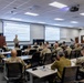 A military briefing or training session is taking place in a modern classroom or conference room, with one uniformed presenter standing at a podium addressing approximately 20-25 seated military personnel. Two projection screens display slides with text related to Army MOS (Military Occupational Specialty) transformation initiatives and Army National Guard topics. The attendees are wearing U.S. Army OCP (Operational Camouflage Pattern) uniforms, with at least one individual in a different olive-drab uniform visible in the foreground.
