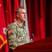 A U.S. Army soldier in OCP (Operational Camouflage Pattern) uniform is speaking at a wooden podium during what appears to be an official military ceremony or briefing. The soldier's name tape reads 'ARMSTRONG' and the uniform displays a U.S. flag patch and what appears to be a Ranger tab on the left shoulder. Multiple military flags and a red curtain backdrop are visible behind the speaker.