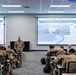 A military briefing is taking place in a modern classroom setting where a uniformed soldier stands at the front presenting to a group of seated military personnel. Two large projection screens display slides titled 'U.S. Army Western Hemisphere Command' featuring maps and strategic information. The audience consists of multiple soldiers in camouflage uniforms seated at individual desks.