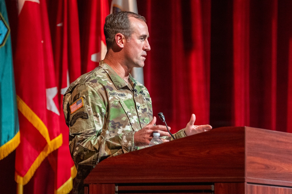 A U.S. Army officer with the name tape 'Armstrong' is speaking at a wooden podium during what appears to be an official military ceremony or briefing. He is wearing Operational Camouflage Pattern (OCP) uniform with a U.S. flag patch and Colonel rank insignia visible. The setting includes multiple military flags and a red curtain backdrop on a stage.