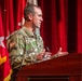 A U.S. Army officer with the name tape 'Armstrong' is speaking at a wooden podium during what appears to be an official military ceremony or briefing. He is wearing Operational Camouflage Pattern (OCP) uniform with a U.S. flag patch and Colonel rank insignia visible. The setting includes multiple military flags and a red curtain backdrop on a stage.