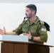 An Israel Defense Forces (IDF) officer is speaking at a wooden podium with a microphone. He is wearing an olive-green IDF uniform with an Israeli flag patch on the right sleeve and a rank insignia on the shoulder. The setting appears to be a formal briefing room or lecture hall.