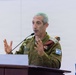 A male military officer in Israeli Defense Forces (IDF) uniform is speaking at a wooden podium with a microphone. He is gesturing with both hands raised, suggesting active engagement with an audience. The setting appears to be an indoor briefing or conference environment.