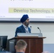 A man wearing a blue turban and blazer is presenting at a wooden podium during a formal conference or briefing. Behind him, a projected slide displays the text 'Develop Technology, Inform R...' dated 15 September 2026, marked 'DISTRIBUTION A: Approved for Public Re...'. A Dell computer monitor and tower are visible in the foreground, and an audience member is partially visible at the bottom of the frame.