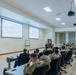 A military briefing or lecture is taking place in a modern conference room, with a civilian presenter standing at a podium addressing a large audience of uniformed military personnel. Two projection screens display slides with organizational charts or diagrams, with the left screen appearing to reference 'Gaza and Lebanon Events' based on partially visible text. The audience is seated at long tables in what appears to be a professional military education or training facility.