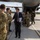 Department of War Delegation visits Poland A civilian official in a dark suit and tie is greeting military personnel in camouflage uniforms on an airport tarmac. The group appears to have just disembarked from a large military transport aircraft visible in the background. The setting suggests an official arrival or diplomatic/military coordination meeting at a military airfield.