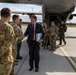 A civilian official in a dark suit and tie is greeting military personnel in camouflage uniforms on an airport tarmac. The group appears to have just disembarked from a large military transport aircraft visible in the background. The setting suggests an official arrival or diplomatic/military coordination meeting at a military airfield.